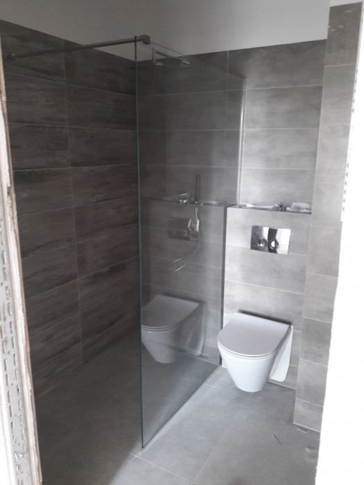 Nowoczesna łazienka z kabiną prysznicową typu walk-in i podwieszanymi toaletami, wykończona szarymi płytkami imitującymi drewno, z armaturą w kolorze chromu.