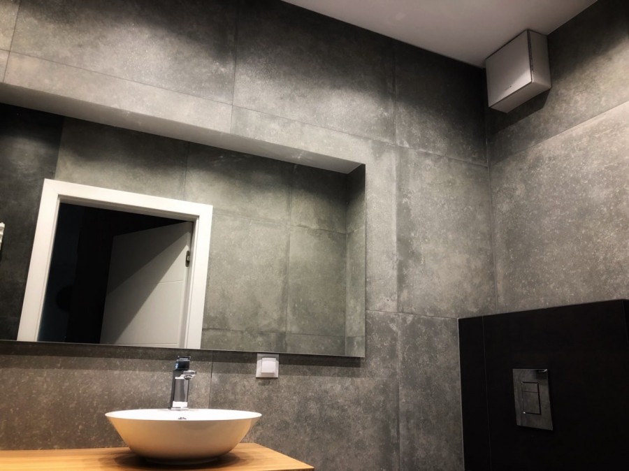 Nowoczesna łazienka z szarymi płytkami imitującymi beton, białą umywalką nablatową na drewnianym blacie, lustrem z podświetleniem i minimalistyczną armaturą.