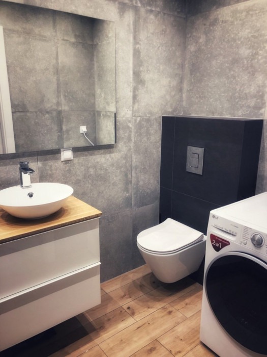 Nowoczesna łazienka z szarymi płytkami imitującymi beton, drewnianą podłogą, umywalką nablatową na szafce z drewnianym blatem, wiszącą toaletą i pralką.