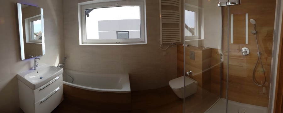 Jasna łazienka z drewnopodobnymi płytkami, kabiną prysznicową ze szklanymi drzwiami, podświetlanym lustrem nad umywalką z szafką, grzejnikiem drabinkowym i wiszącą toaletą.