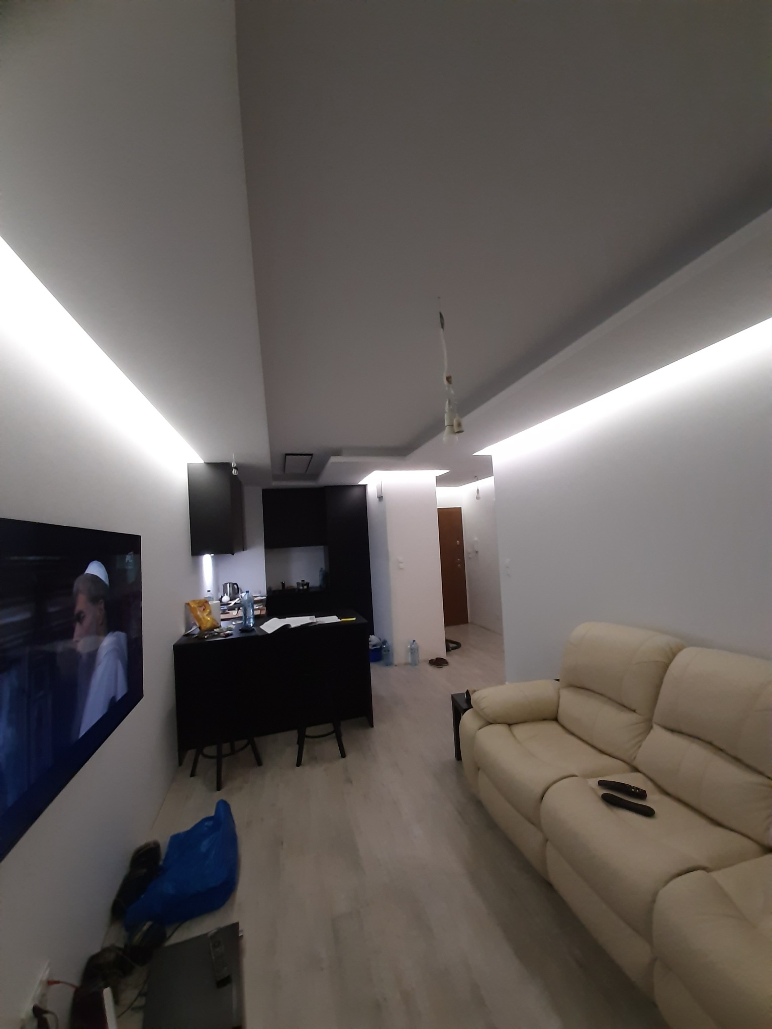 Nowoczesne wnętrze mieszkania po remoncie: białe ściany z oświetleniem LED, jasna podłoga, beżowa sofa, czarna zabudowa kuchenna z barkiem i telewizor na ścianie.