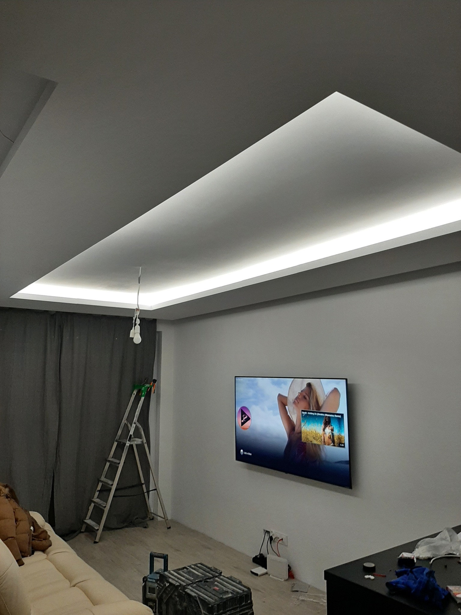 Nowoczesny sufit napinany z oświetleniem LED w salonie, widoczna drabina i narzędzia sugerujące niedawny montaż.