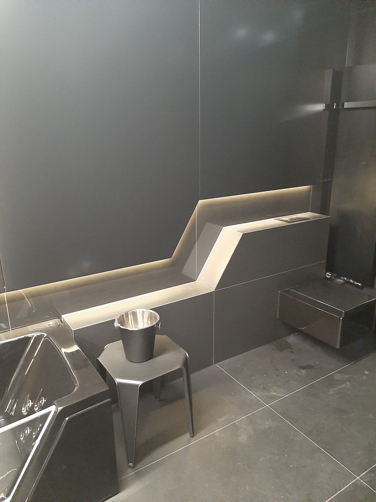 Minimalistyczna łazienka w odcieniach szarości z podświetleniem LED, czarna wanna, metalowe wiaderko na stołku, płytki wielkoformatowe na ścianach i podłodze.