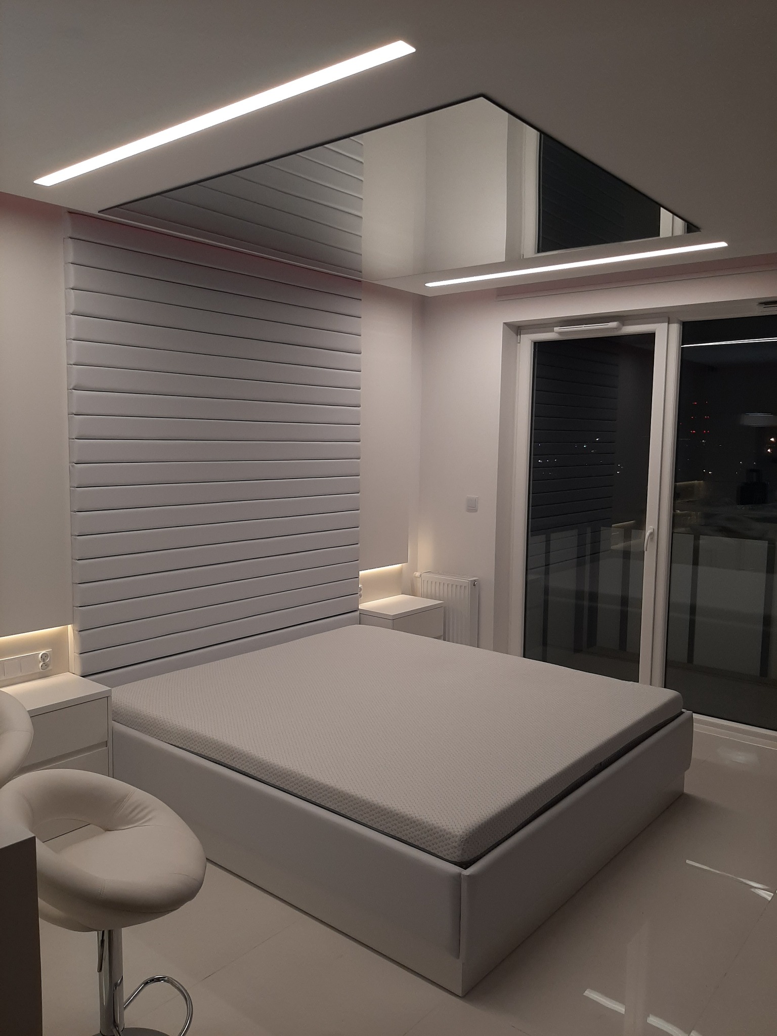 Nowoczesna sypialnia z białym łóżkiem, panelem ściennym za wezgłowiem i podwieszanym sufitem z oświetleniem LED, widok na okno balkonowe.