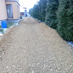 Przygotowanie podłoża z kruszywa pod drogę dojazdową w Olkuszu, widoczne obrzeża z ziemi i rząd wysokich tuj.