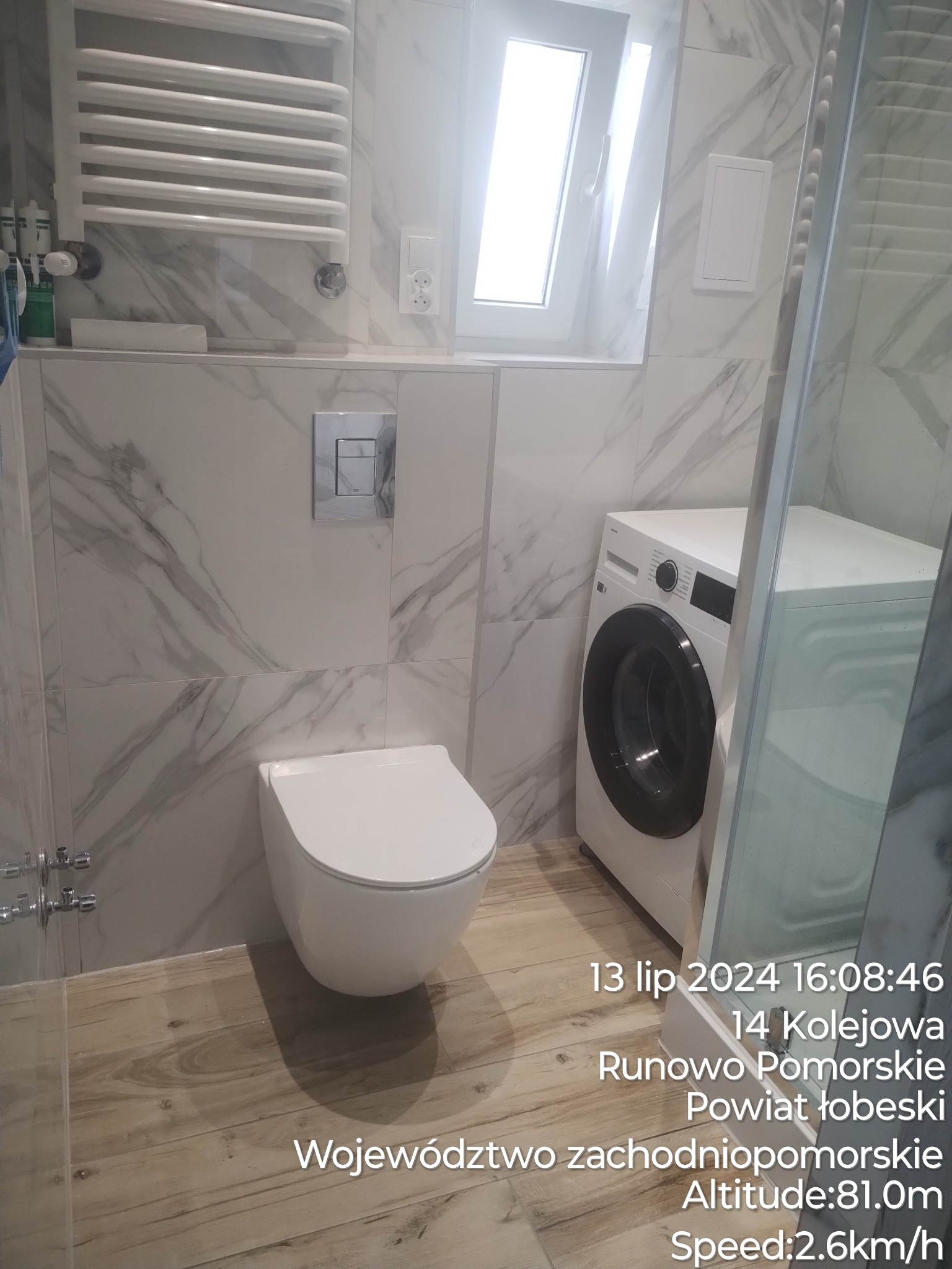 Nowoczesna łazienka z białymi marmurkowymi płytkami, wiszącą toaletą, grzejnikiem drabinkowym, pralką i drewnopodobną podłogą, sfotografowana z perspektywy kabiny prysznicowej.