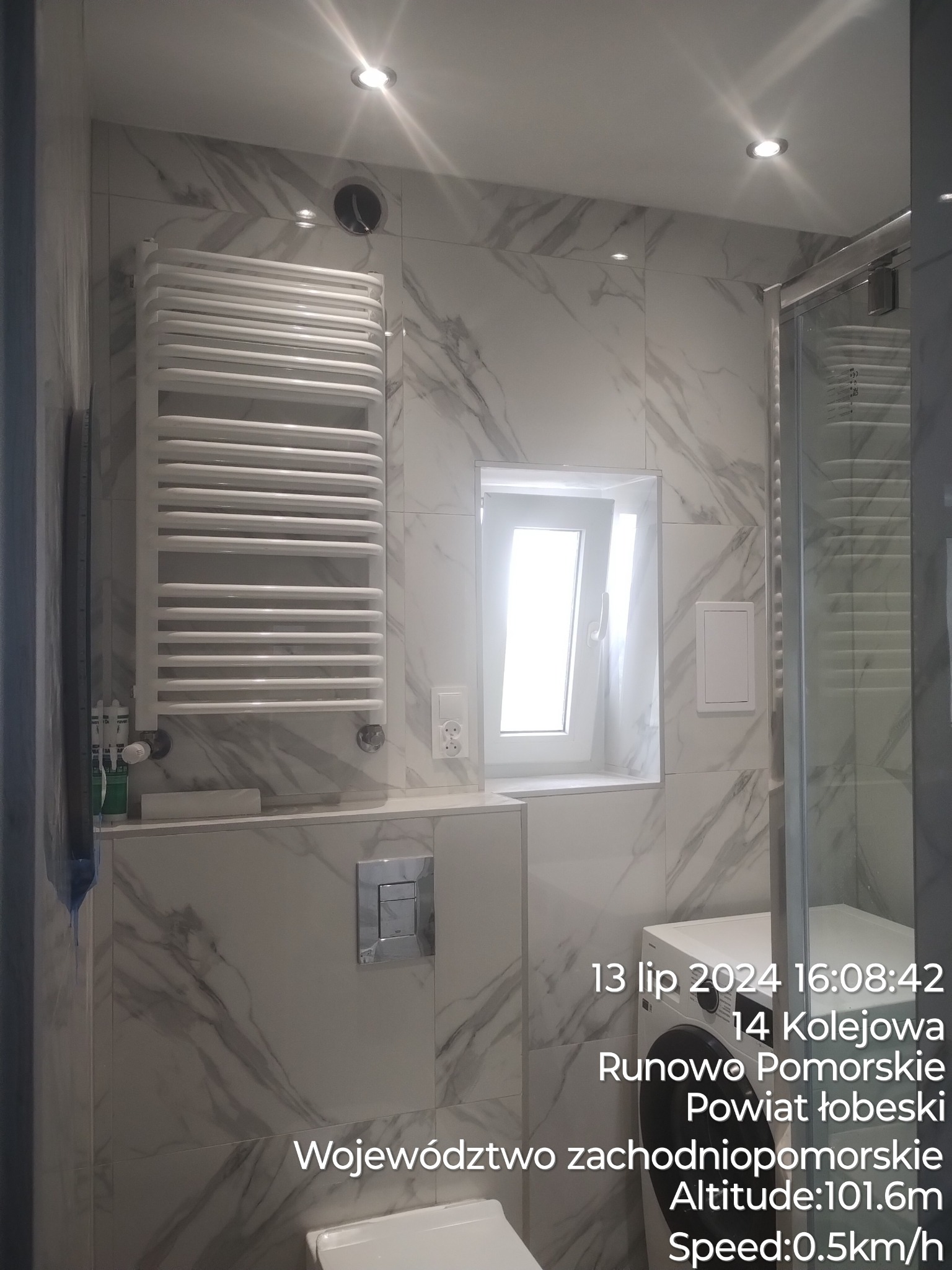 Nowoczesna łazienka z marmurowymi płytkami, grzejnikiem drabinkowym, oknem wnękowym i kabiną prysznicową, widoczne detale wykończenia.