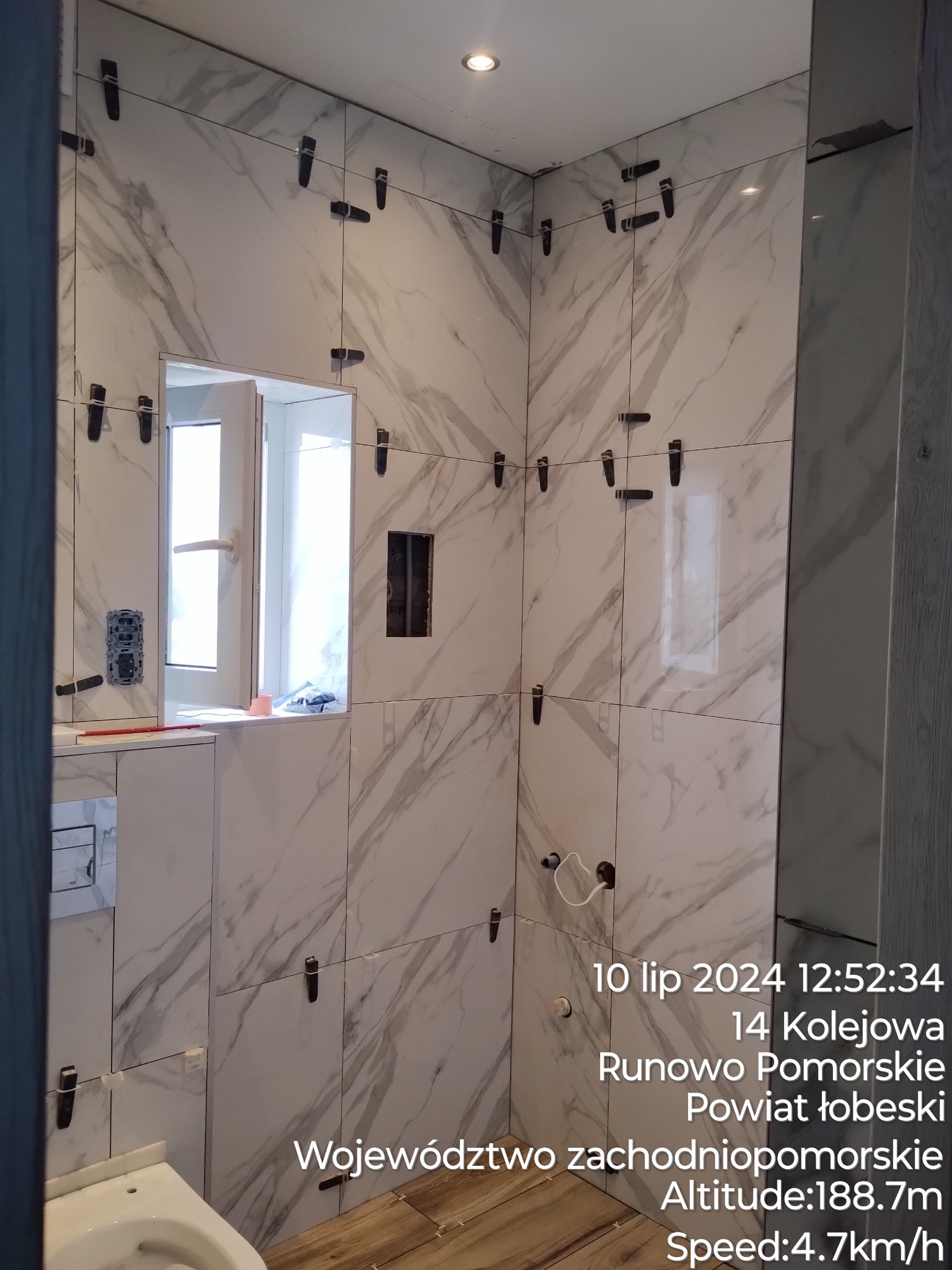 W trakcie układania płytek imitujących marmur w łazience, widoczne czarne klipsy poziomujące, częściowo widoczna toaleta i drewnopodobna podłoga, okno w tle.