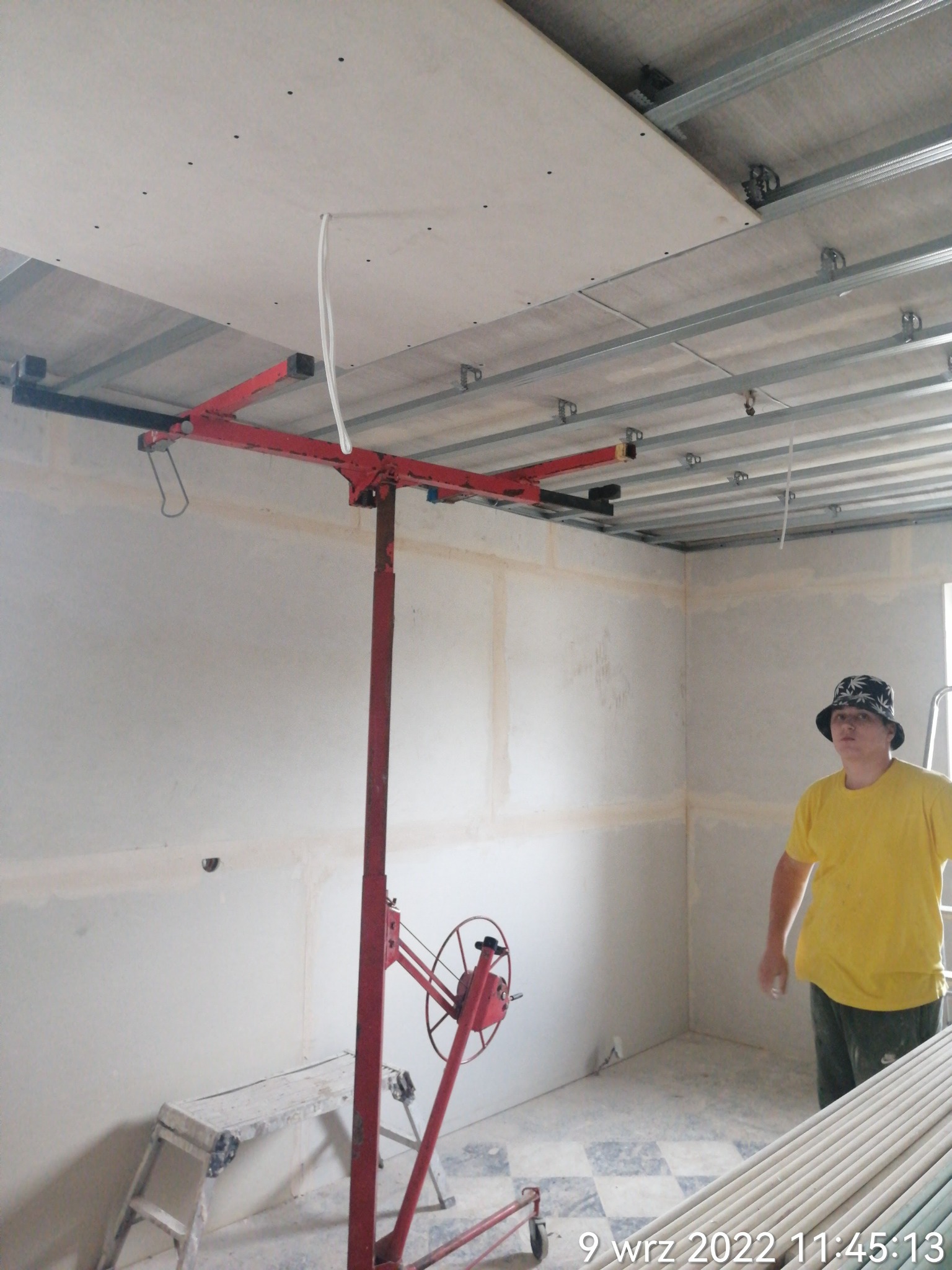 Montaż płyty gipsowo-kartonowej na suficie za pomocą podnośnika; widoczna metalowa konstrukcja nośna, częściowo zamontowana płyta, biały przewód elektryczny oraz pracownik w żółtej koszulce...