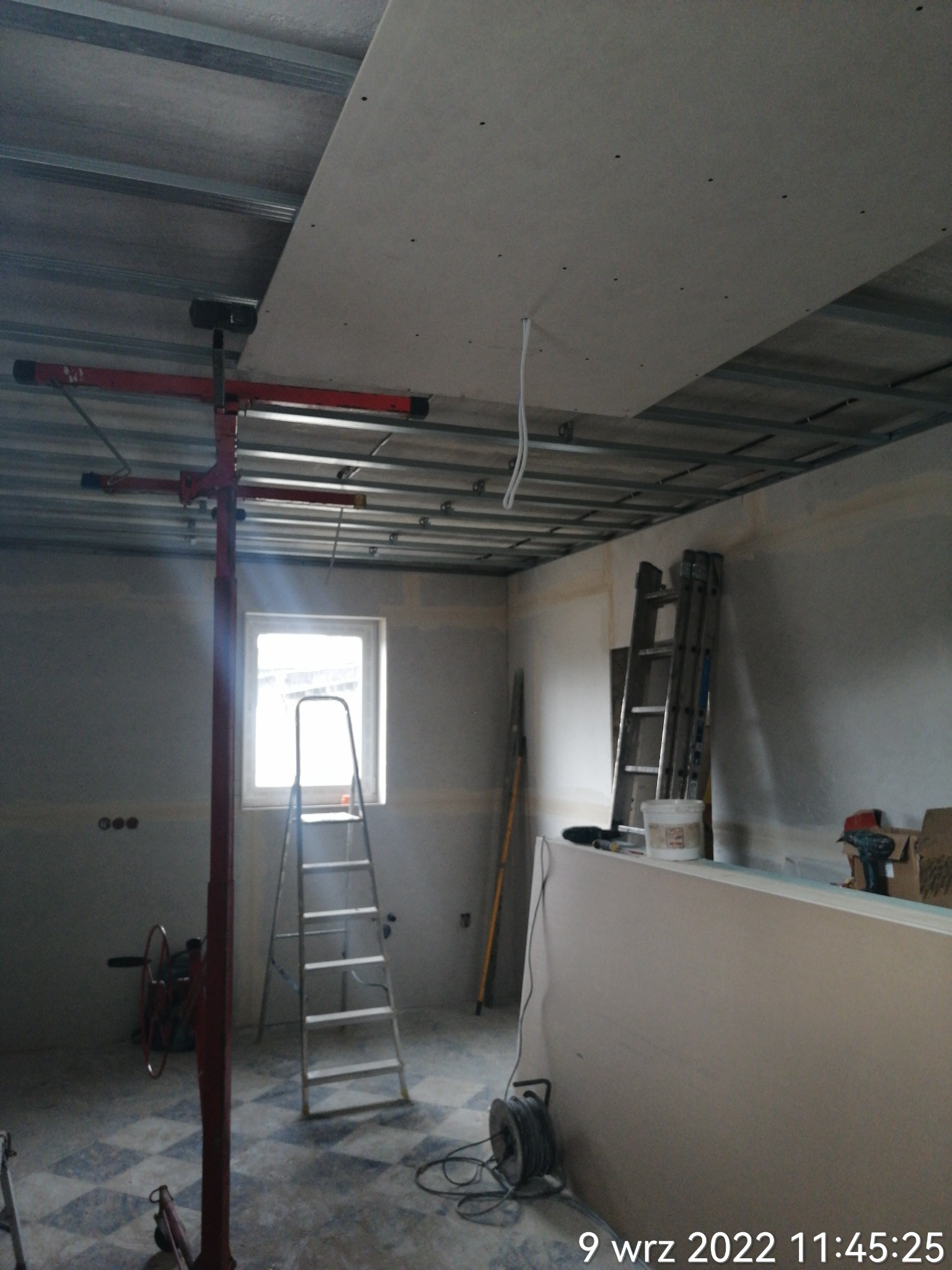 Montaż płyty gipsowo-kartonowej na suficie podwieszanym, widoczny podnośnik do płyt, metalowa konstrukcja sufitu, drabina oparta o ścianę, narzędzia i materiały budowlane w pomieszczeniu w trakcie...