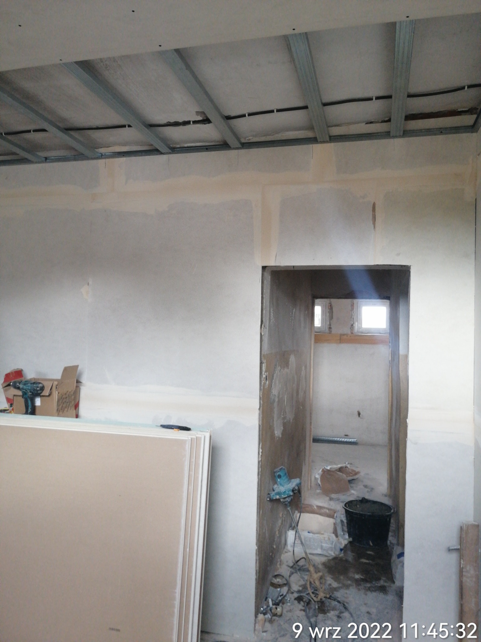 Wnętrze w trakcie prac wykończeniowych: płyty gipsowo-kartonowe oparte o ścianę, widoczna konstrukcja metalowa sufitu podwieszanego, ściany częściowo pokryte gładzią, bałagan budowlany w przejściu...