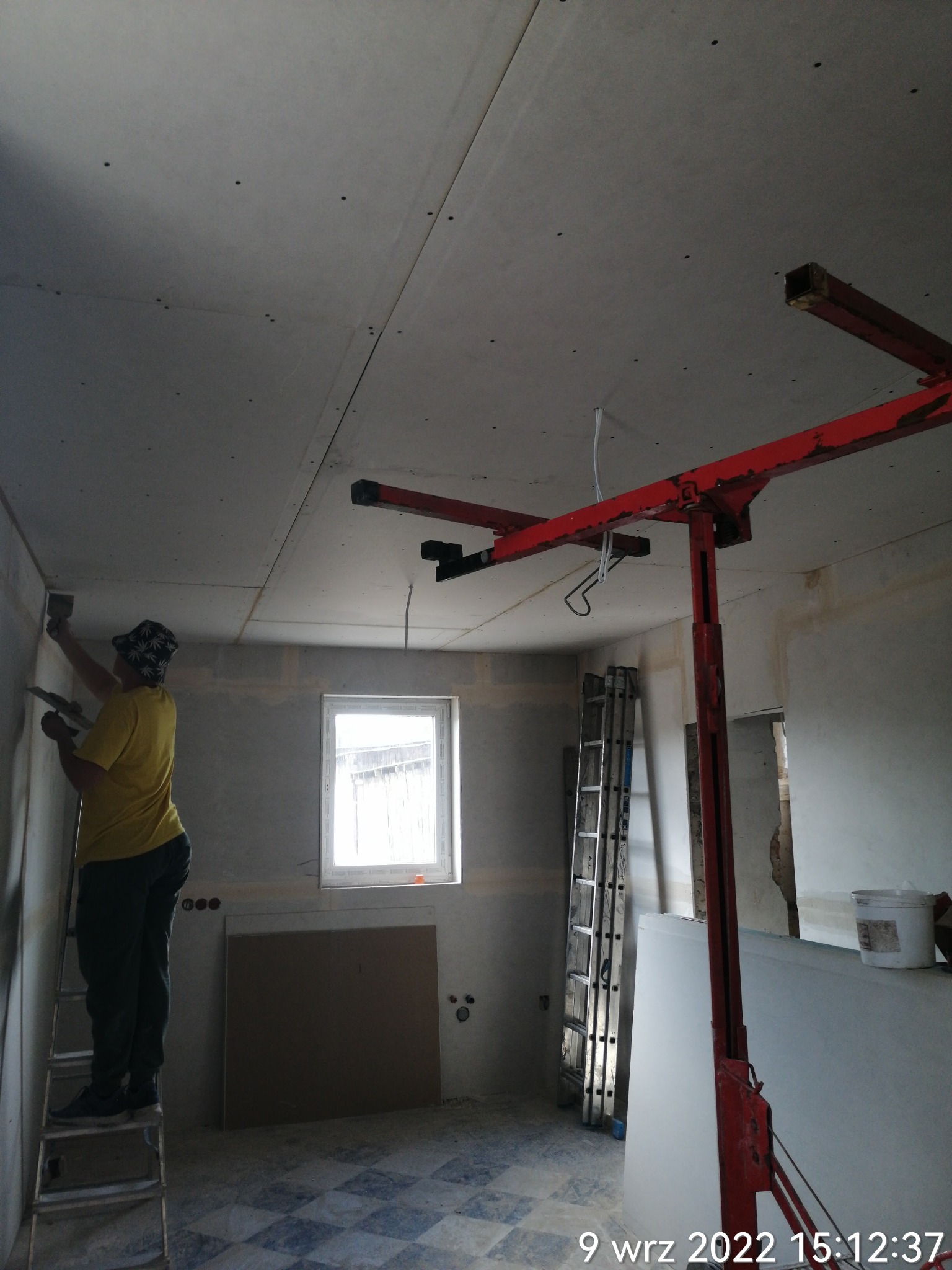 Pracownik na drabinie szpachluje łączenie płyt gipsowo-kartonowych na ścianie, widoczny podnośnik do płyt, sufit z zamontowanymi płytami, okno i fragment podłogi w kratkę.