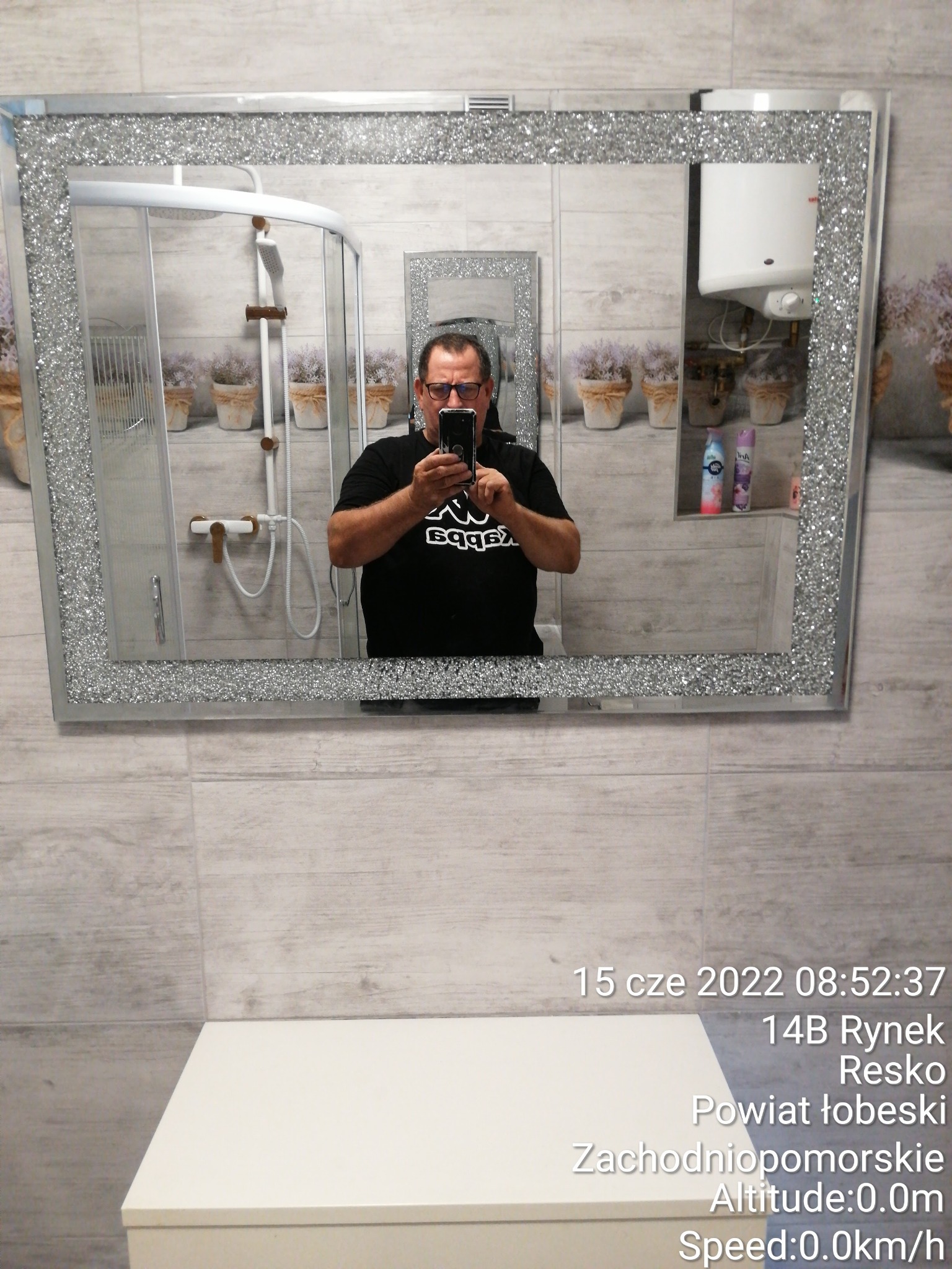 Odbicie w lustrze z brokatową ramą, ukazujące łazienkę z kabiną prysznicową, bojlerem i półką z dekoracyjnymi kwiatami w doniczkach, fotograf robiący selfie w lustrze.