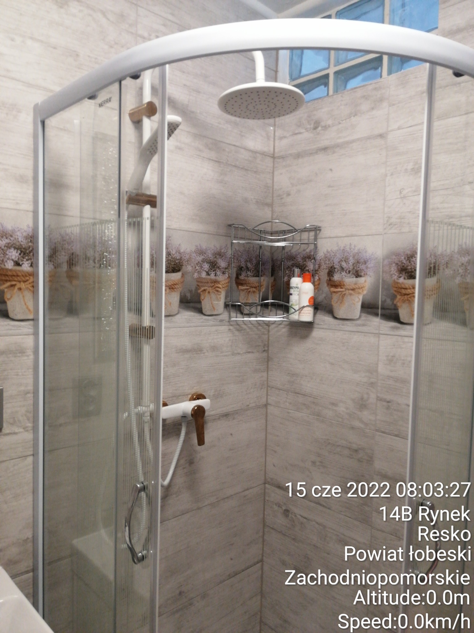 Kabina prysznicowa z deszczownicą i panelem natryskowym, widoczne płytki imitujące drewno oraz półka z kosmetykami i dekoracyjne doniczki z lawendą.