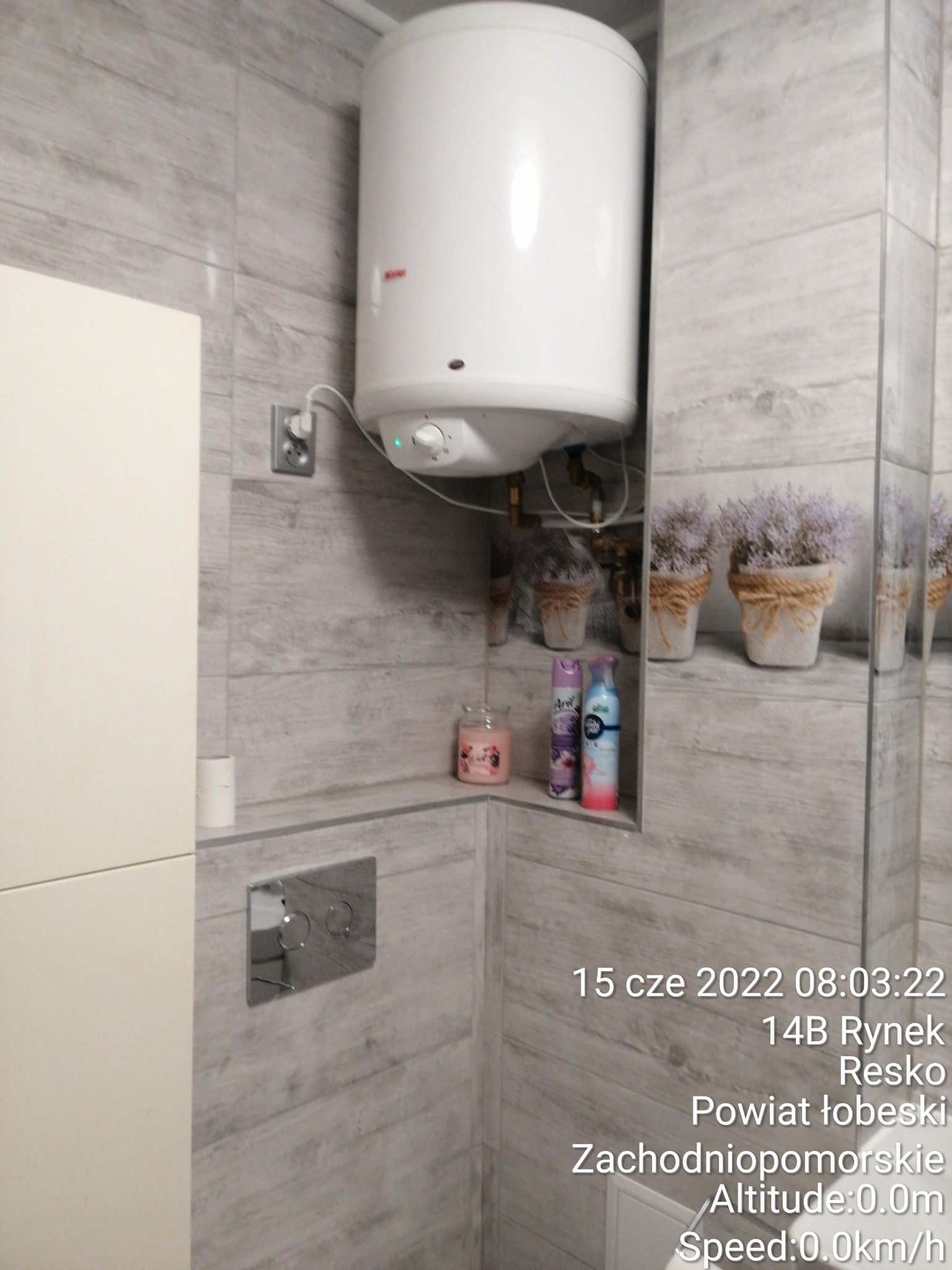 Bojler elektryczny zamontowany na szarej ścianie z płytek imitujących drewno w wyremontowanej łazience, widoczna również spłuczka podtynkowa, półka z dekoracjami i odbicie w lustrze.