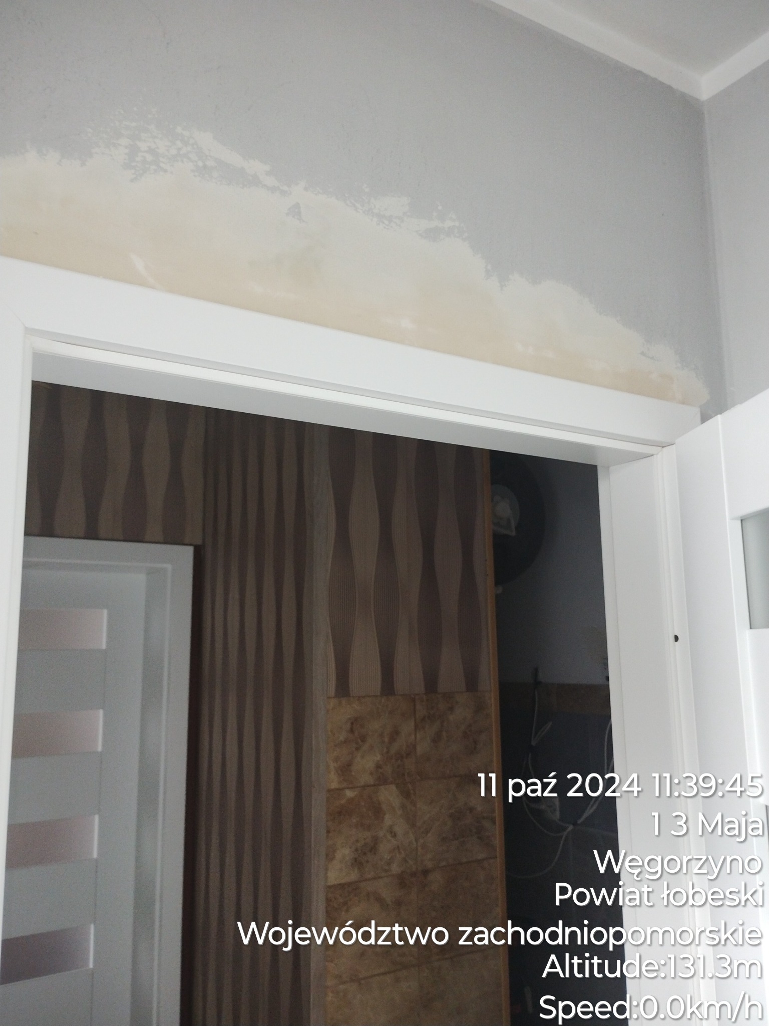 Ściana po usunięciu tapety, widoczne ślady kleju i przygotowanie do malowania, biała ościeżnica drzwiowa, w tle fragment wzorzystej tapety.