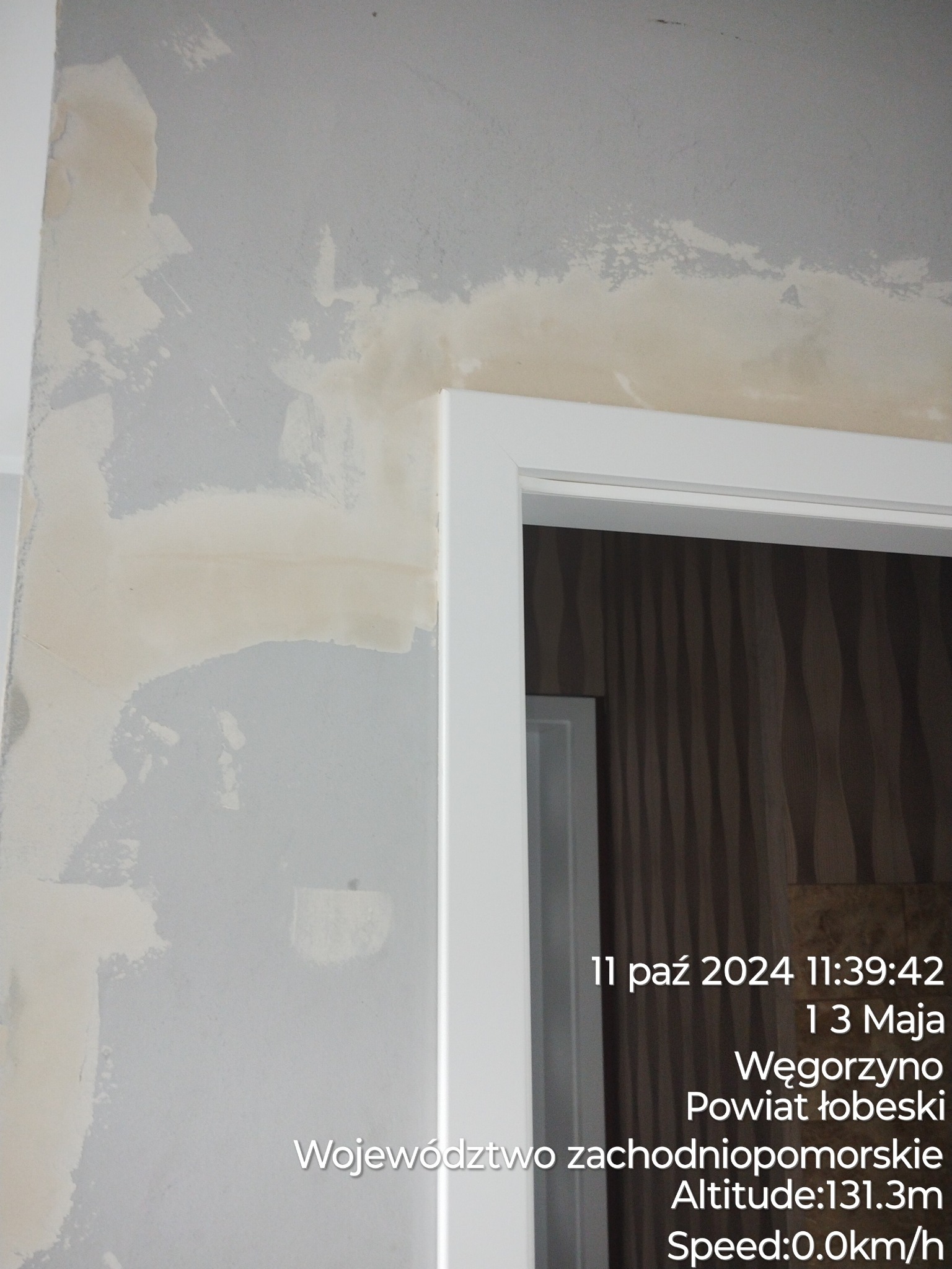 Fragment ściany w trakcie szpachlowania, widoczne białe ślady po gładzi szpachlowej na szarej ścianie, z prawej strony biała ościeżnica drzwiowa, w tle tapeta z pionowymi wzorami.