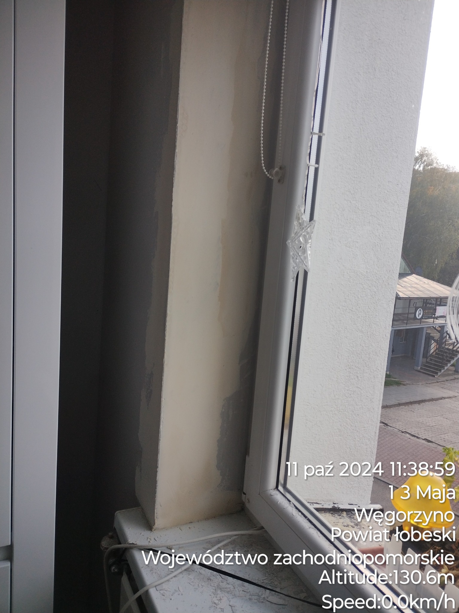 Niedokończone gładzie na ścianie przy oknie, widoczne ślady po szpachlowaniu, białe okno PCV z roletą sznurkową, na parapecie grzejnik.