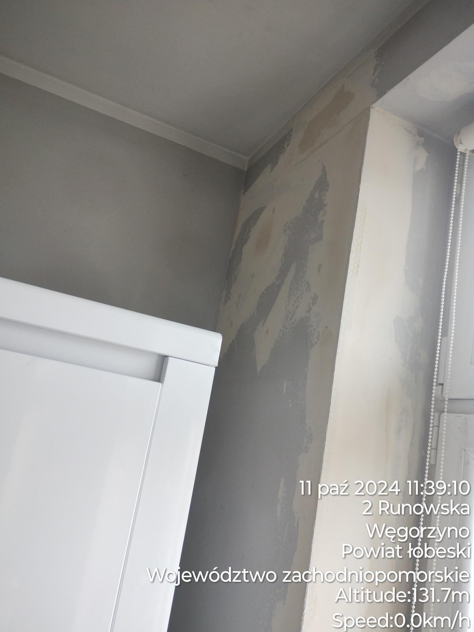 Przygotowanie ściany pod malowanie: zaszpachlowana ściana z widocznymi nierównościami i śladami szpachli, biała roleta okienna w rogu, fragment białego mebla przy ścianie.