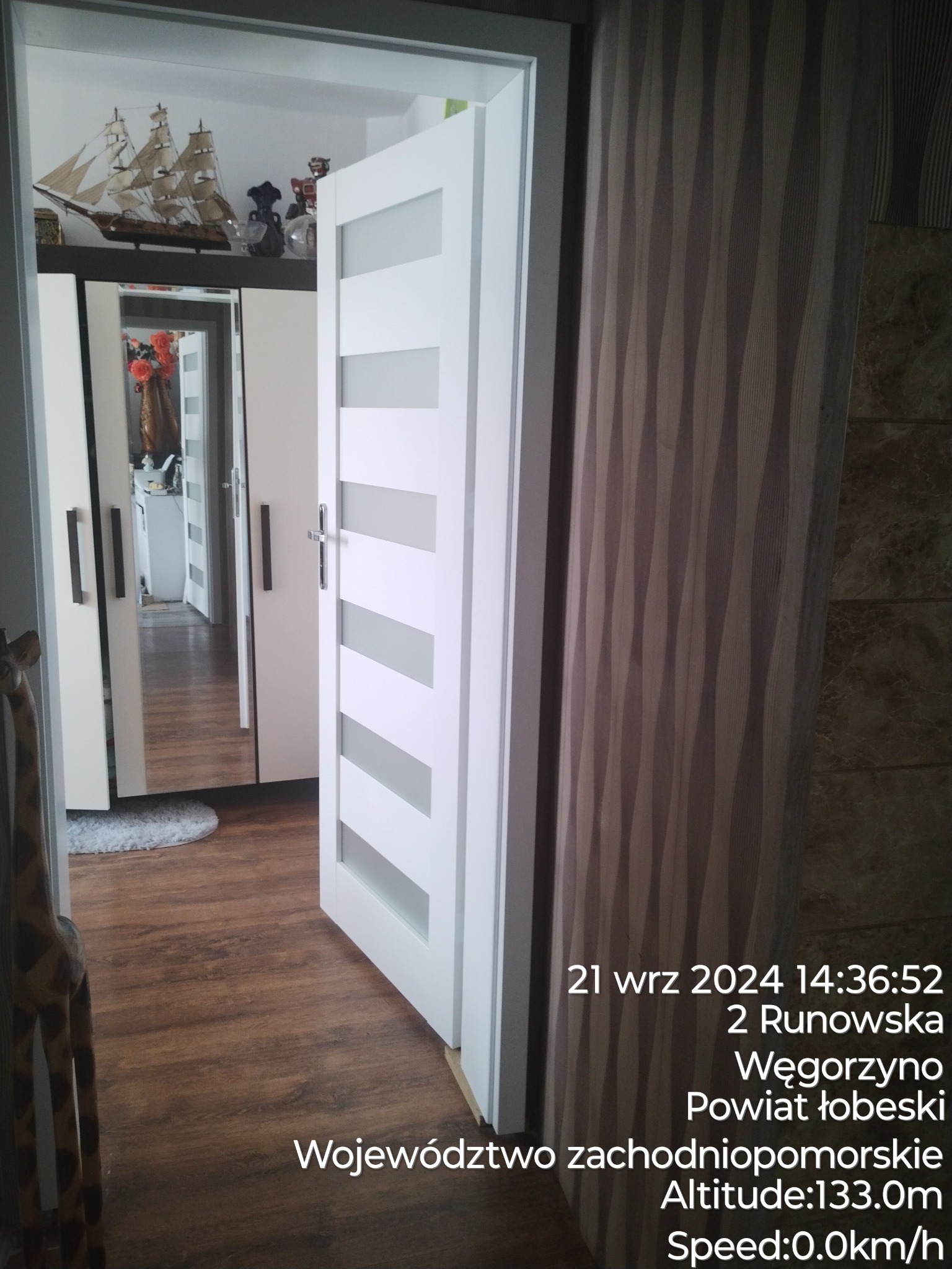 Otwarta biała wewnętrzna drzwi z poziomymi szarymi paskami, widok na lustrzaną szafę i dekoracje w głębi mieszkania, drewniana podłoga.