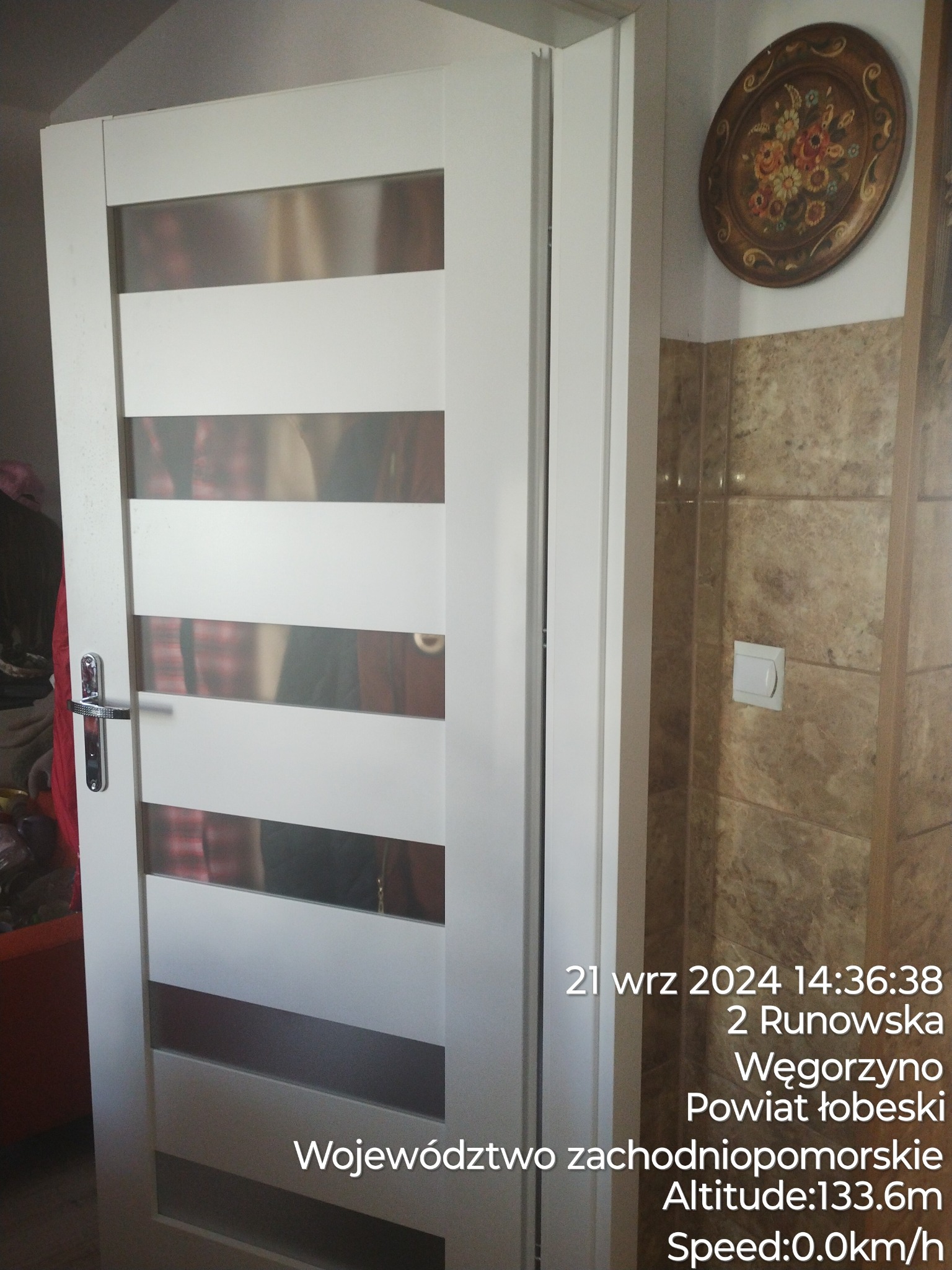 Białe drzwi wewnętrzne z poziomymi, matowymi przeszkleniami, srebrna klamka, na ścianie dekoracyjny talerz i brązowe płytki.