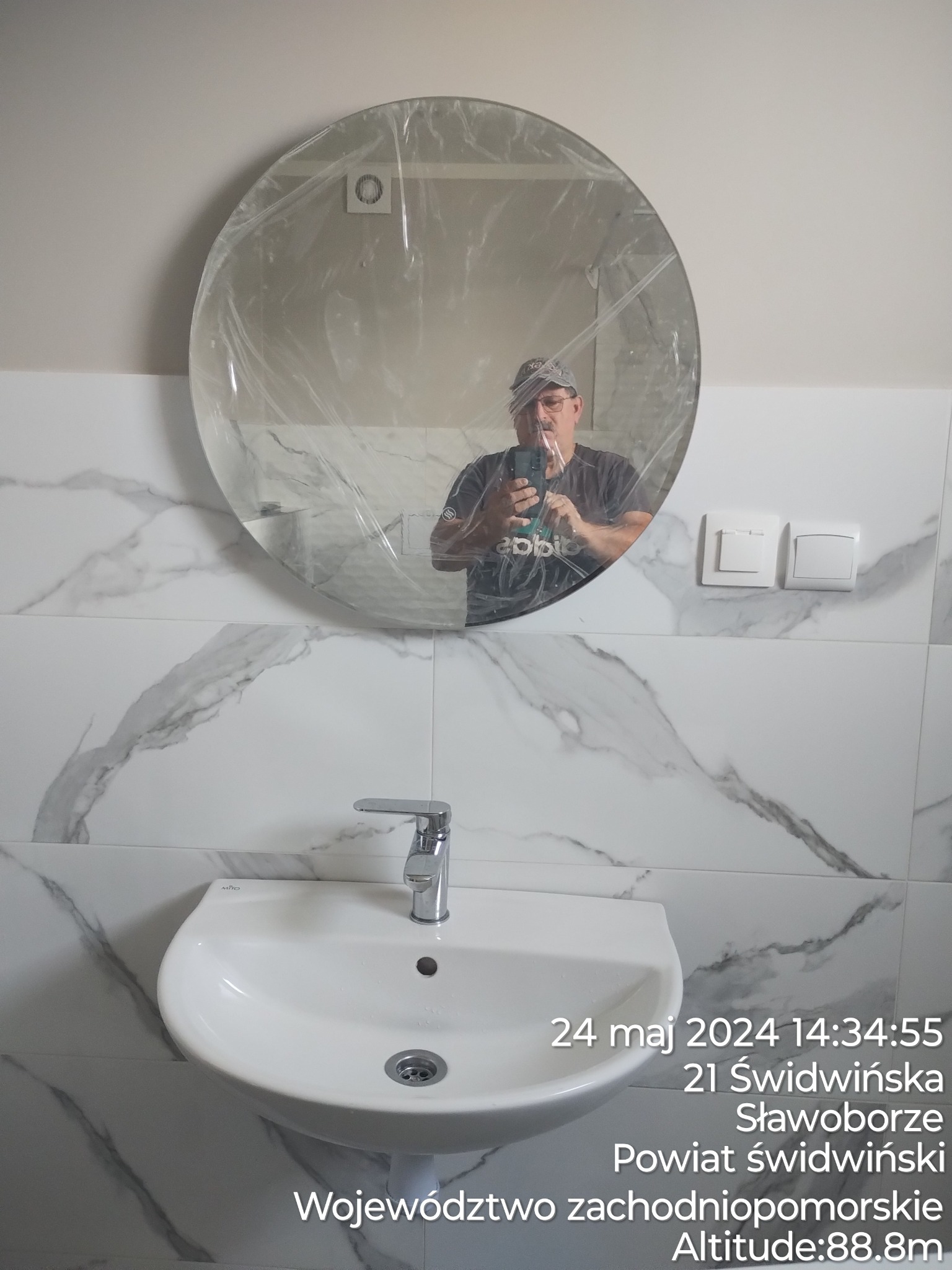 Okrągłe lustro w folii ochronnej nad białą umywalką z chromowaną baterią, marmurowe płytki na ścianie, odbicie fotografa w lustrze.