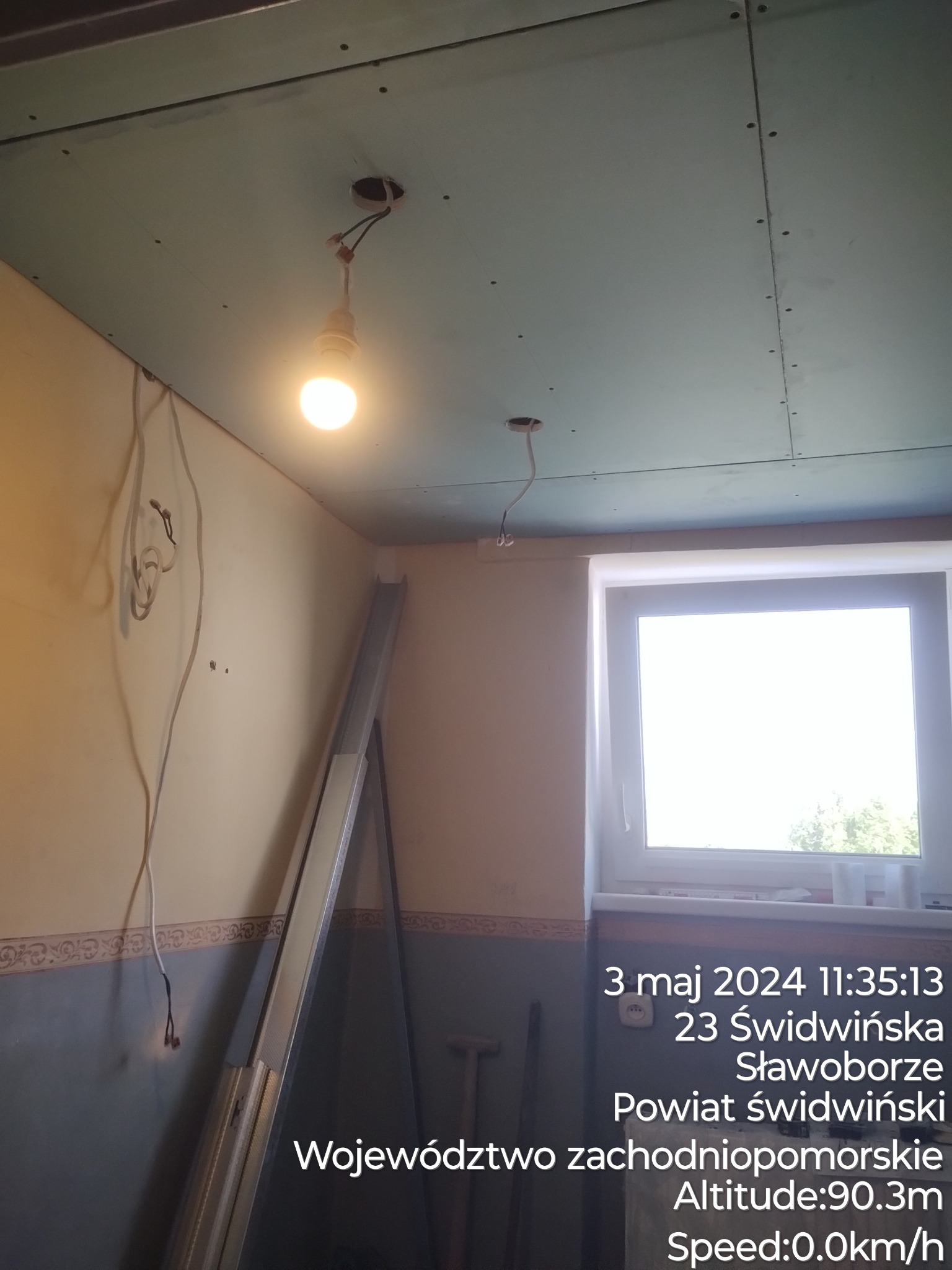 Poddasze w trakcie prac wykończeniowych: zamontowane płyty gipsowo-kartonowe na suficie, widoczne kable elektryczne i tymczasowe oświetlenie żarówką, ściany częściowo pomalowane, okno w tle.