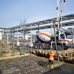 Wylewanie betonu na zbrojenie fundamentu z pompy betonomieszarki, pracownik w żółtych kaloszach i pomarańczowej kamizelce, w tle instalacja przemysłowa.