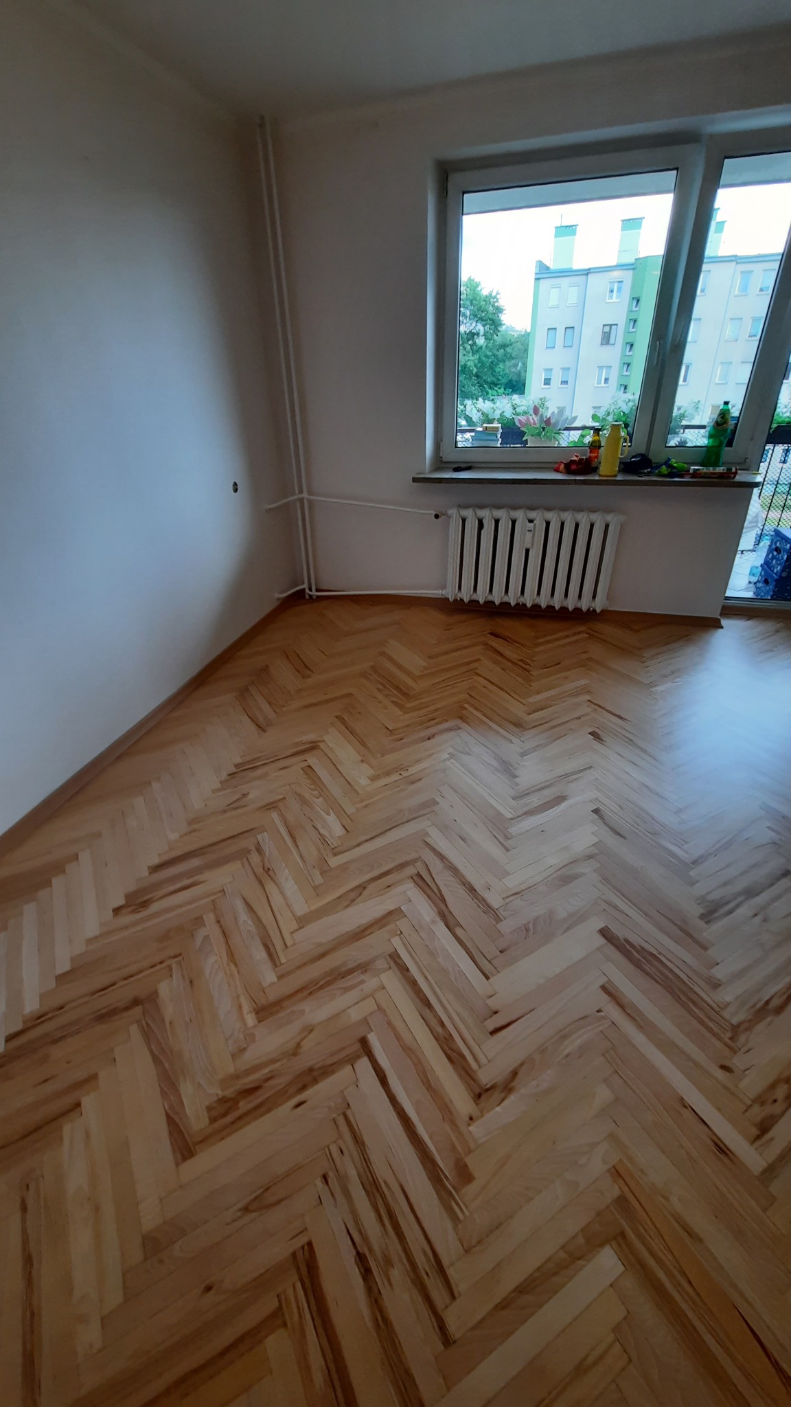 Pokój po remoncie z drewnianą podłogą w jodełkę, biały grzejnik pod oknem z widokiem na blok mieszkalny.