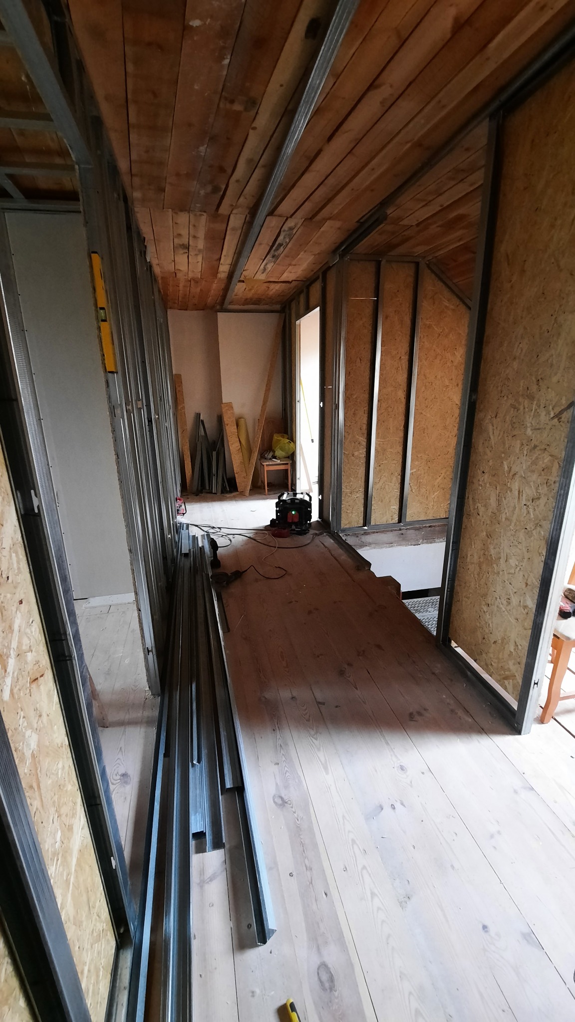 Konstrukcja szkieletowa ścian działowych z metalowych profili, widoczne drewniane deski sufitowe i podłogowe, poziomica na ścianie, narzędzia i materiały budowlane w tle.