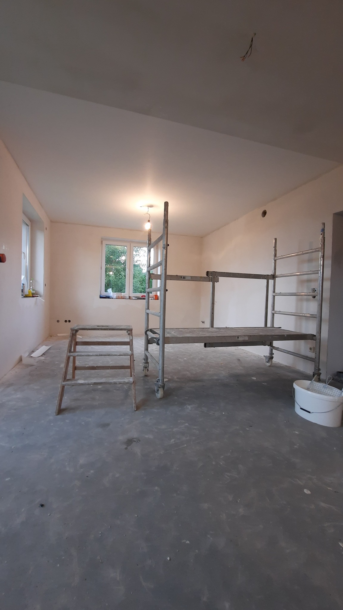 Wnętrze pokoju w trakcie remontu, widoczne rusztowanie, drabina malarska i wiadro z farbą, ściany świeżo pomalowane na jasny kolor.