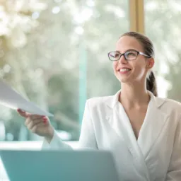Uśmiechnięta kobieta w białym żakiecie i okularach odbiera dokument od osoby z poza kadru, z laptopem na biurku i rozmyte tło.