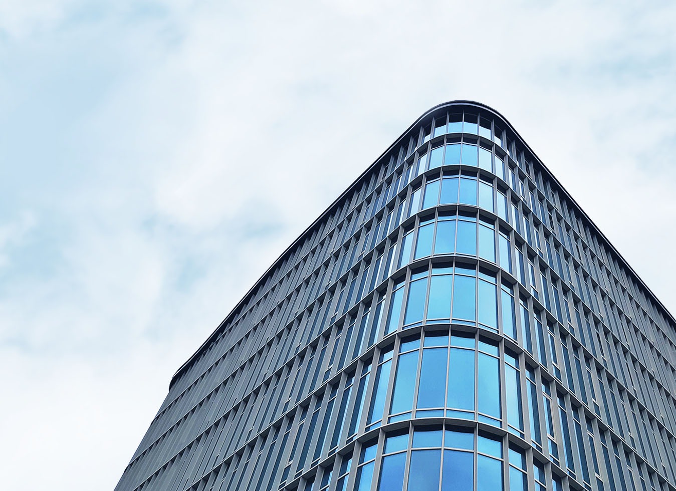 Niski kąt widzenia na narożnik nowoczesnego biurowca z zaokrąglonymi oknami, na tle pochmurnego nieba.