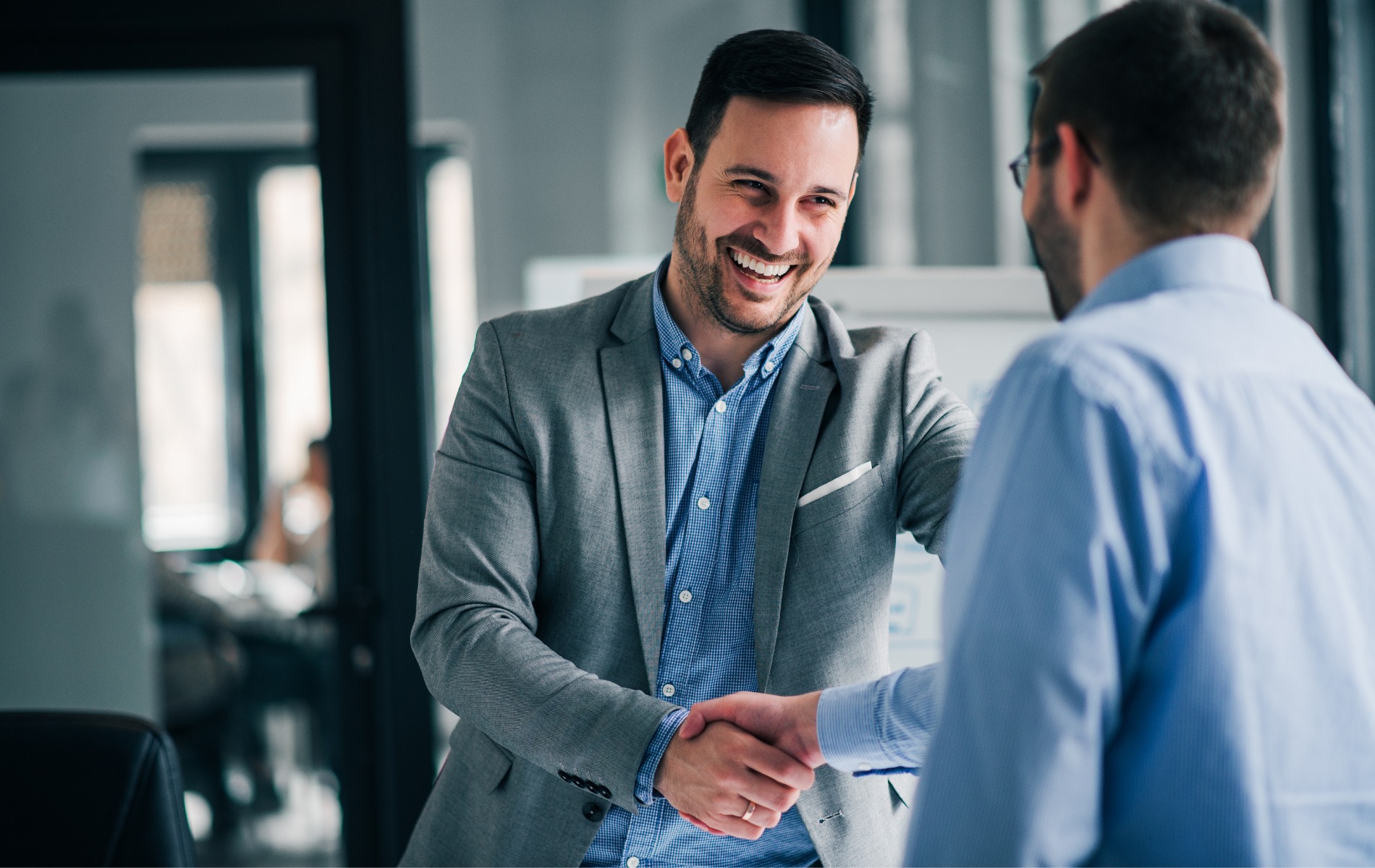 Uśmiechnięty mężczyzna w szarej marynarce i niebieskiej koszuli ściska dłoń innej osobie w jasnym biurze, sugerując zawarcie umowy.