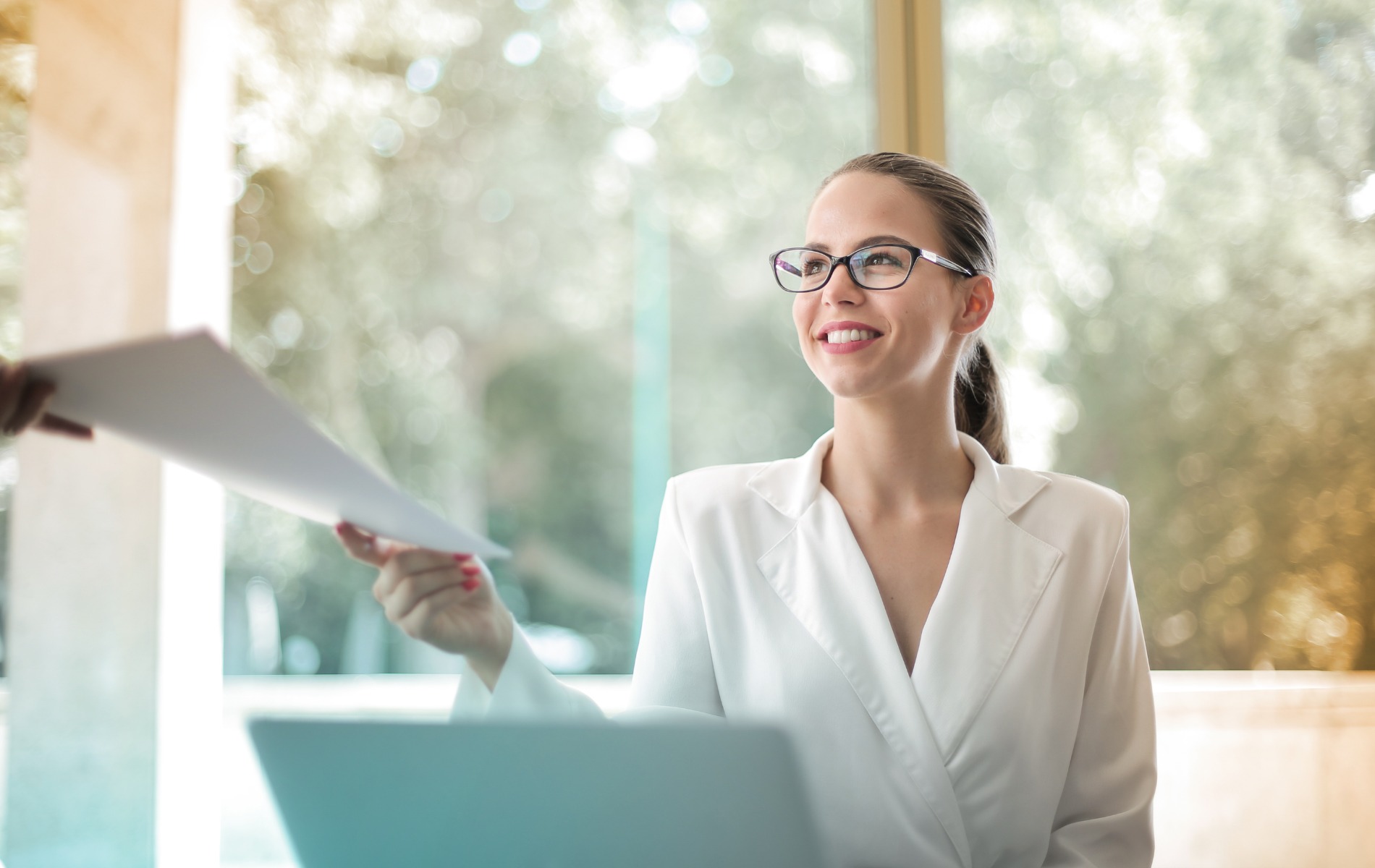 Uśmiechnięta kobieta w białym żakiecie i okularach odbiera dokument od osoby z poza kadru, z laptopem na biurku i rozmyte tło.