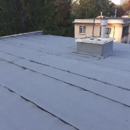 Płaski dach pokryty papą termozgrzewalną z widocznymi łączeniami, komin w tle, widok z góry na budynek otoczony drzewami.