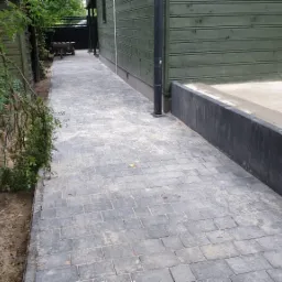 Ułożona szara kostka brukowa tworząca ścieżkę obok zielonego domu z drewnianą elewacją, widoczne drzewa i krzewy po jednej stronie ścieżki.