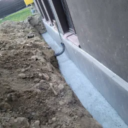 Wykonana izolacja pionowa fundamentu z widocznym wykopem, gruntem i wystającą rurą karbowaną przy oknie.