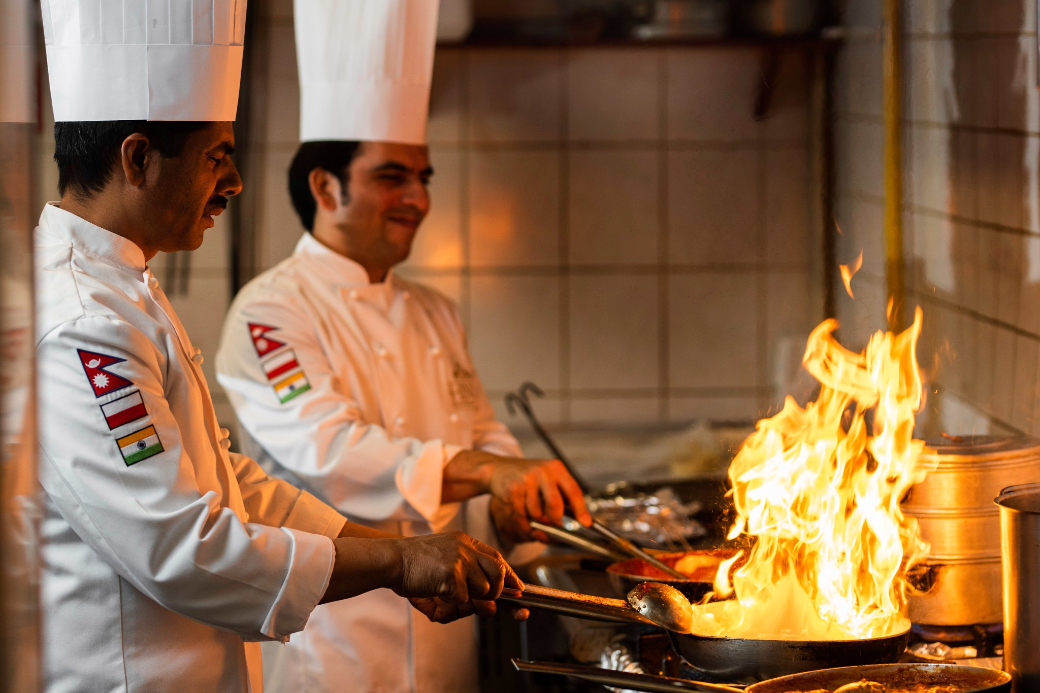 Dwóch kucharzy w białych uniformach z naszywkami flag państwowych na ramionach, przygotowuje danie na patelni, z której wydobywają się płomienie ognia.