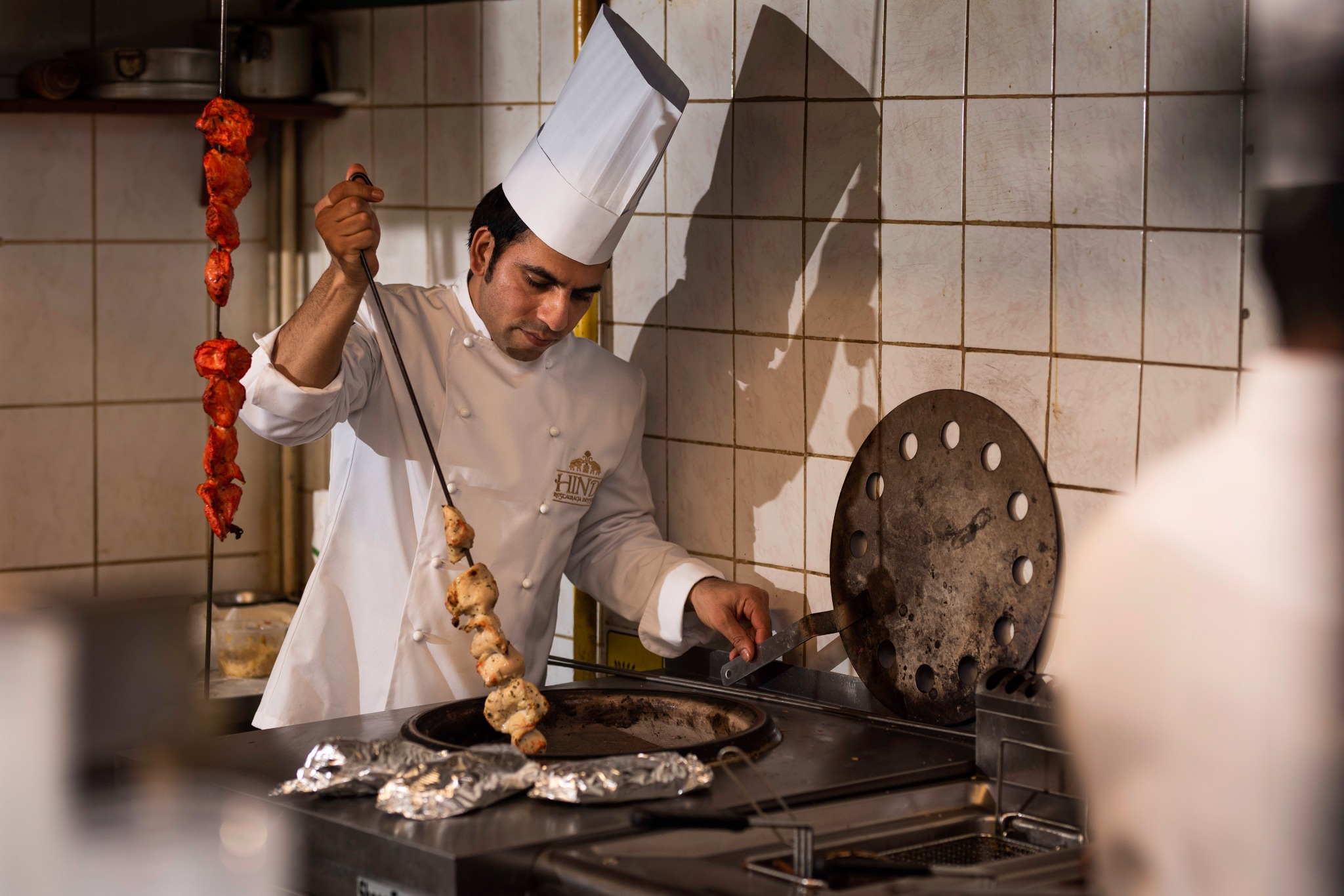 Kucharz w białym uniformie i wysokiej czapce wkłada szaszłyk z kurczaka do pieca tandoor, w tle widoczne szaszłyki z czerwonym mięsem, logo restauracji na uniformie.