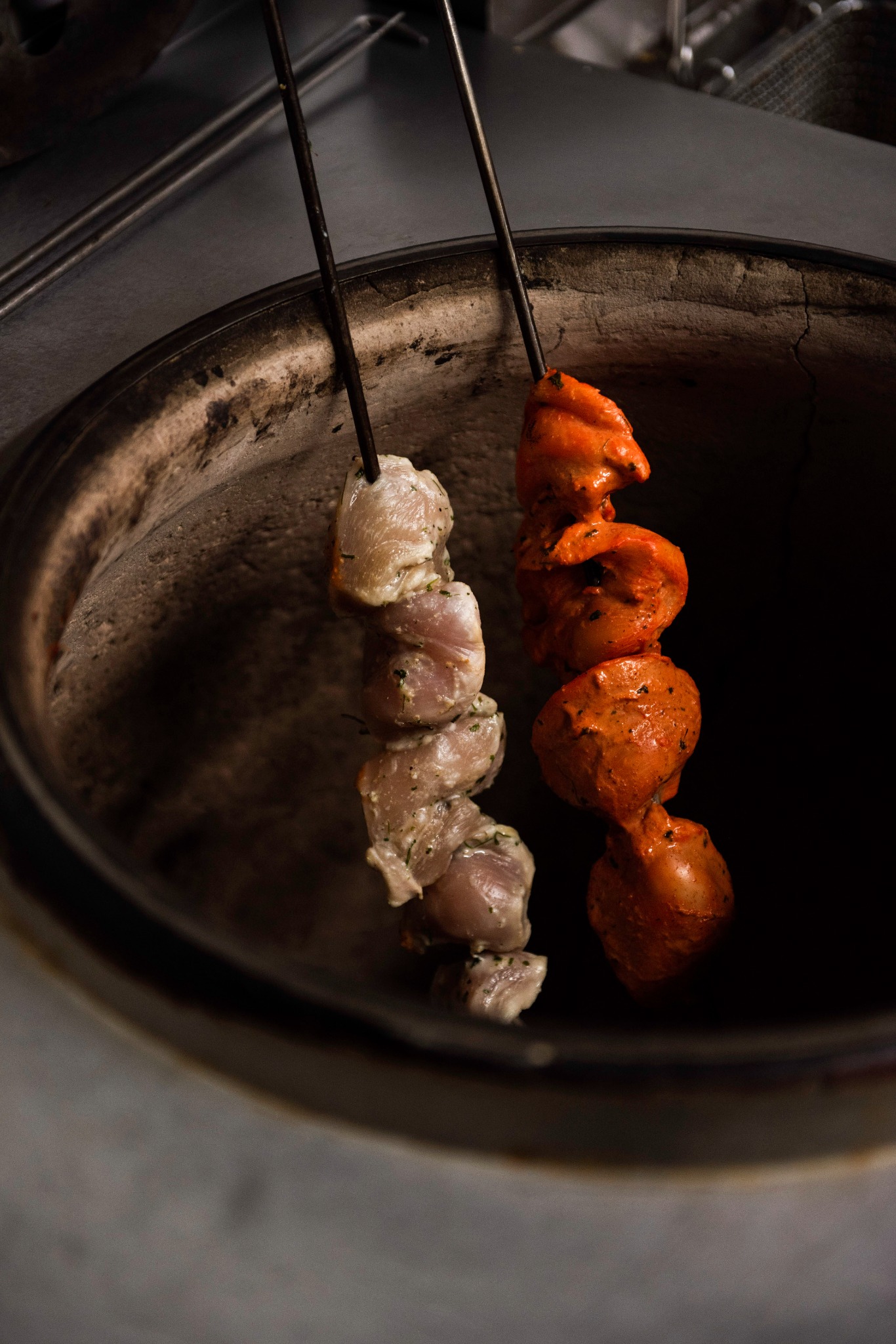 Dwa szaszłyki z marynowanego mięsa (jeden jasny, drugi pomarańczowy) przygotowywane w tradycyjnym piecu tandoor.