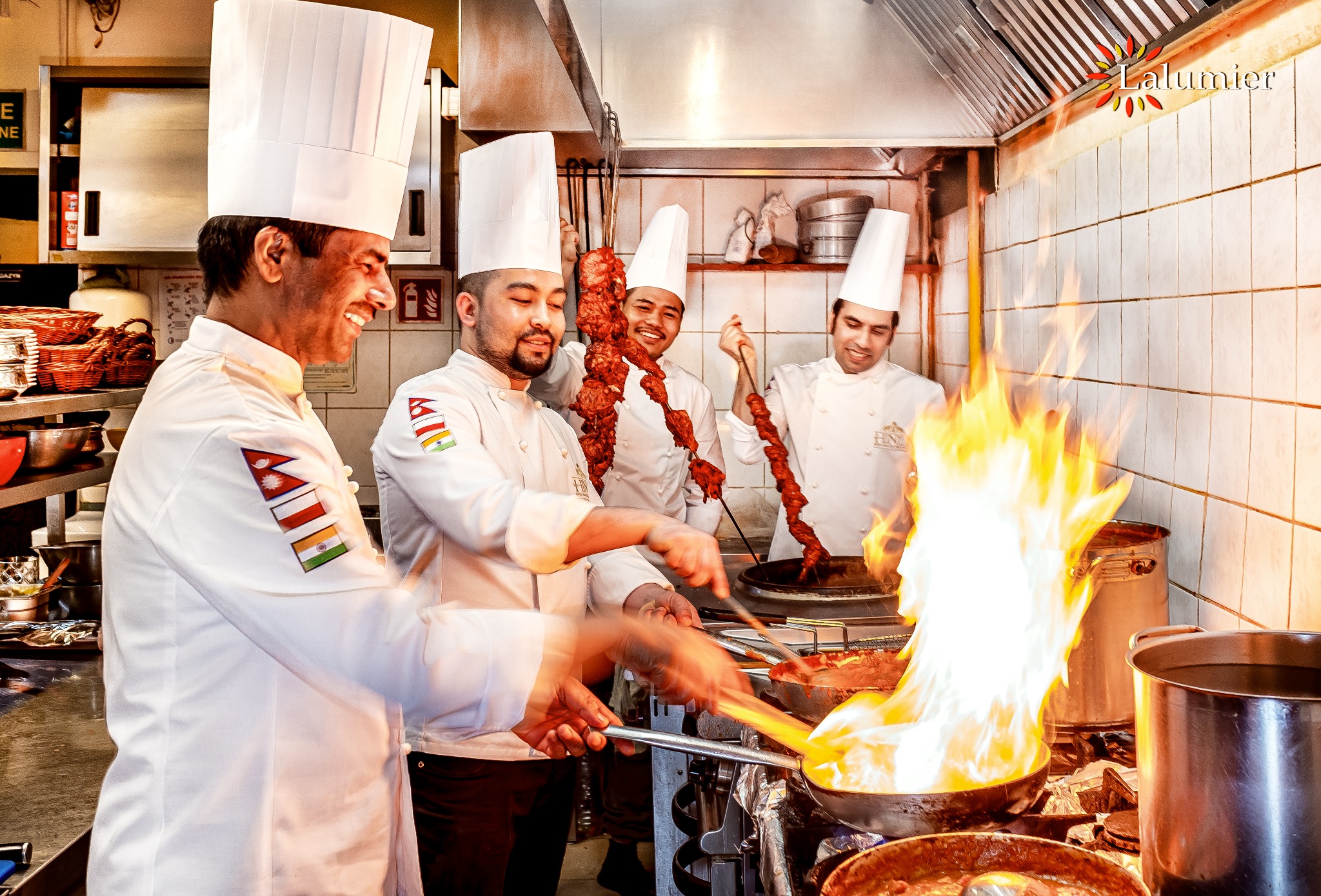 Szczęśliwi kucharze w kuchni, w białych uniformach z flagami Nepalu i Indii na ramionach, przygotowują szaszłyki i dania w płomieniach, w tle metalowe garnki i przybory kuchenne.