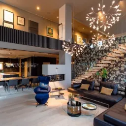 Przestronne, dwupoziomowe wnętrze z otwartą kuchnią, jadalnią i salonem. W centrum salonu znajduje się czarna skórzana sofa narożna, szklany stolik kawowy i designerski fotel w kolorze granatowym.