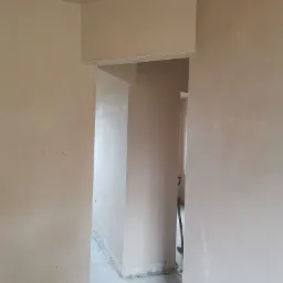 Świeżo nałożony, jasny tynk na ścianach i suficie w przejściu między pomieszczeniami, widoczne resztki materiału na podłodze i narzędzie w tle.