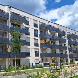 MzGlass - Nowoczesny budynek mieszkalny z balkonami zabezpieczonymi ażurowymi balustradami w kolorze grafitowym, widok z poziomu zadbanego dziedzińca z placem zabaw.