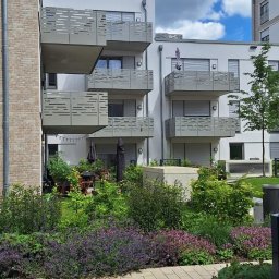 MzGlass - Nowoczesne balkony z ażurowymi osłonami w bloku mieszkalnym, widok z poziomu chodnika w otoczeniu zieleni.