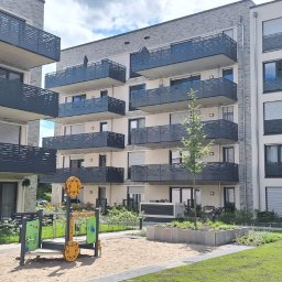 MzGlass - Nowoczesny apartamentowiec z balkonami zabezpieczonymi ciemnymi, ażurowymi balustradami, widok na plac zabaw i zadbany trawnik.