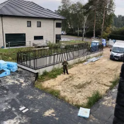 Nowoczesny dom z jasnej cegły z czarną, metalową balustradą na tarasie, widok z góry na plac budowy z pracownikiem w odzieży roboczej.