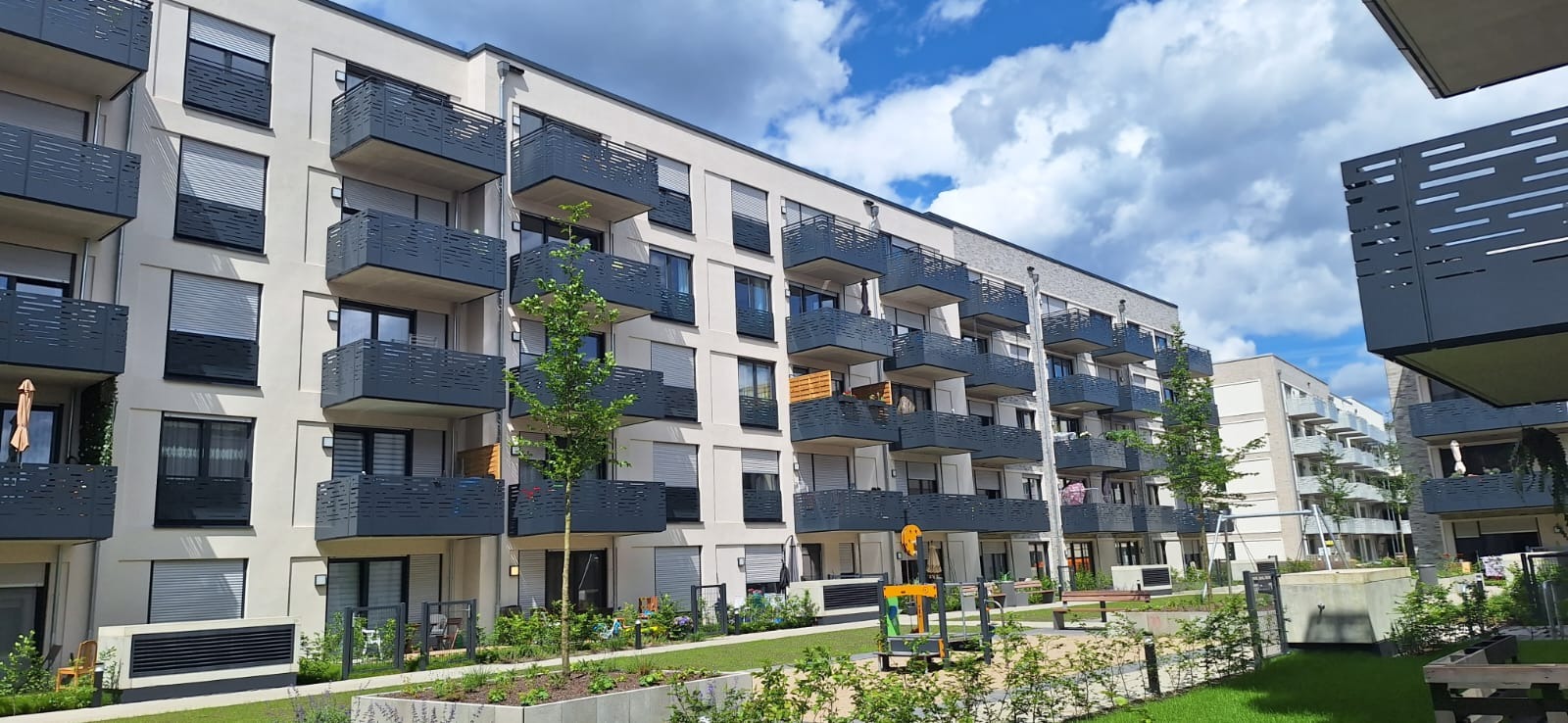 Nowoczesny budynek mieszkalny z balkonami zabezpieczonymi ażurowymi balustradami w kolorze grafitowym, widok z poziomu zadbanego dziedzińca z placem zabaw.