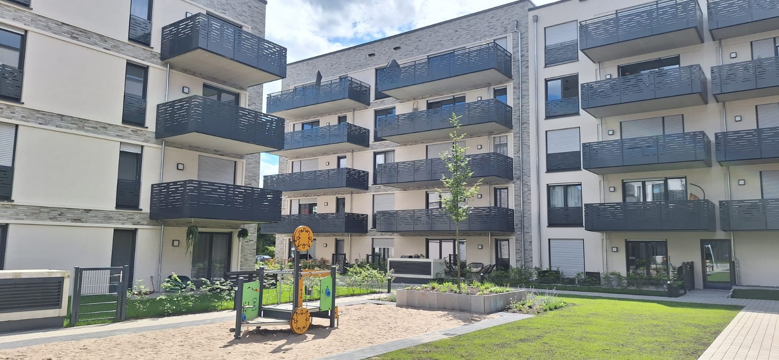 Nowoczesny apartamentowiec z balkonami zabezpieczonymi ciemnymi, ażurowymi balustradami, widok na plac zabaw i zadbany trawnik.